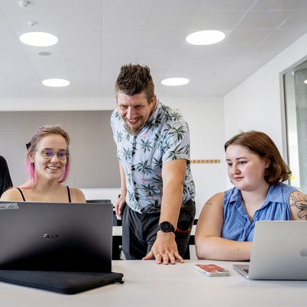 Studerende der sidder og smiler, og underviser der står bøjet hen over bordet.